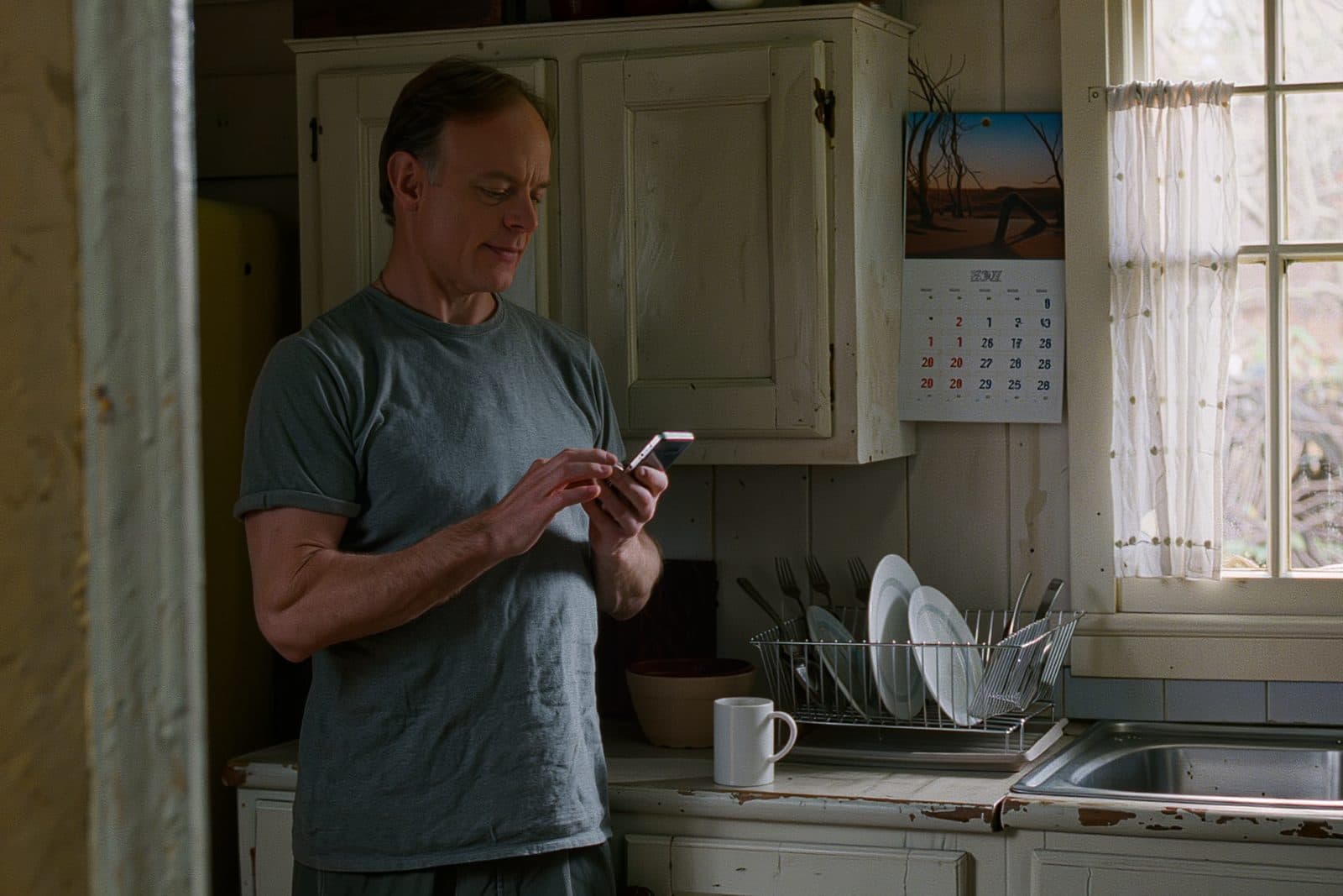A man in his mid forties reading on his phone at a kitchen counter early in the morning.