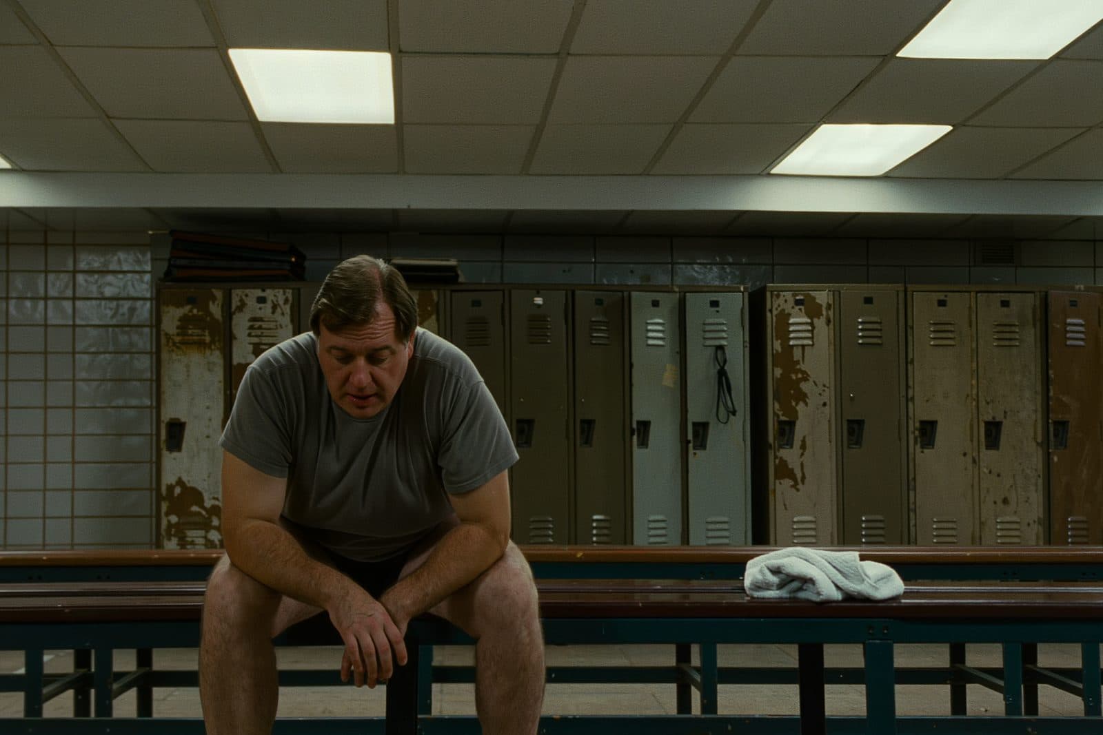 A man in his early forties in an ordinary gym changing room, sitting on a bench putting on a trainer.