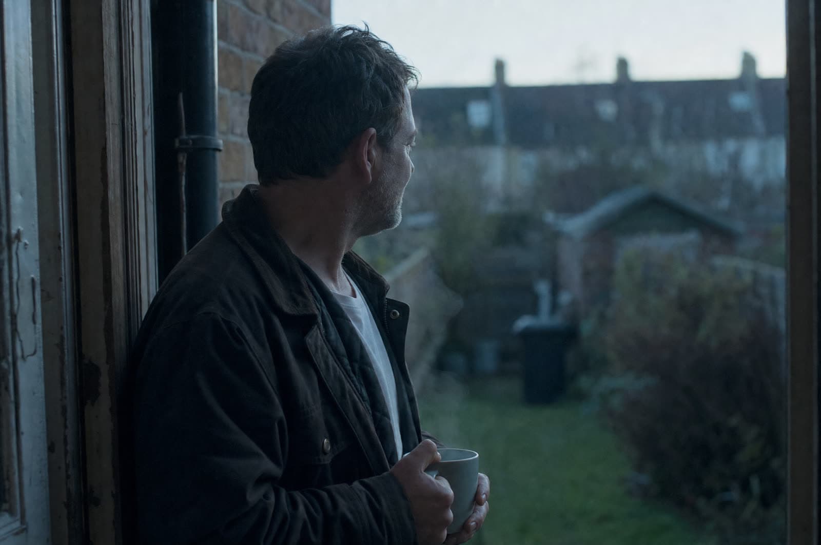 A man in his late forties standing in a back doorway at dawn with a mug of tea, looking out over a terraced garden.